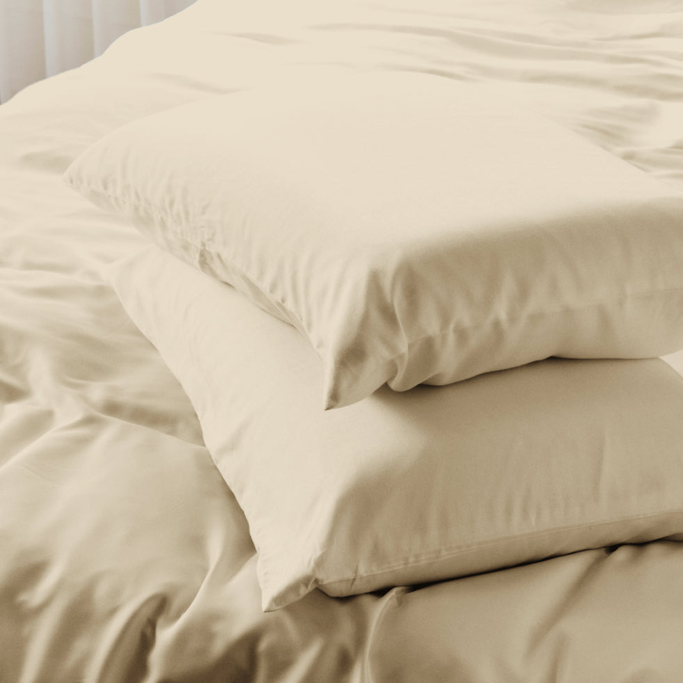A stack of two AURTRS driftwood pillows on a bed with matching cream bedding, shown in a softly lit bedroom setting.